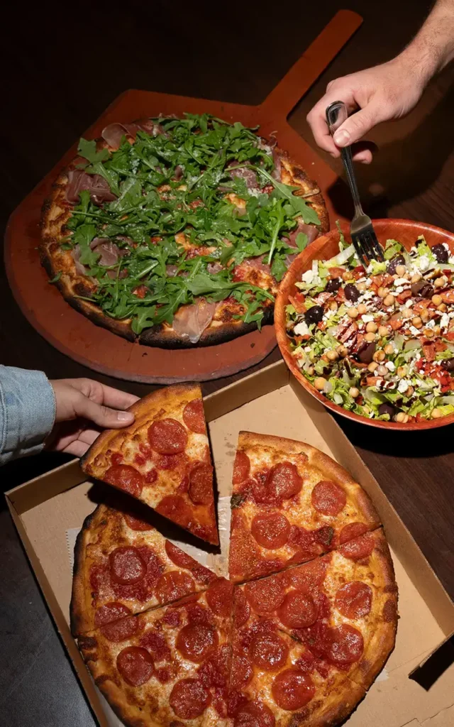 A coal fired pizza topped with prosciutto and arugula beside a loaded salad at Anthony's Coal Fired Pizza in a warm dine-in setting.