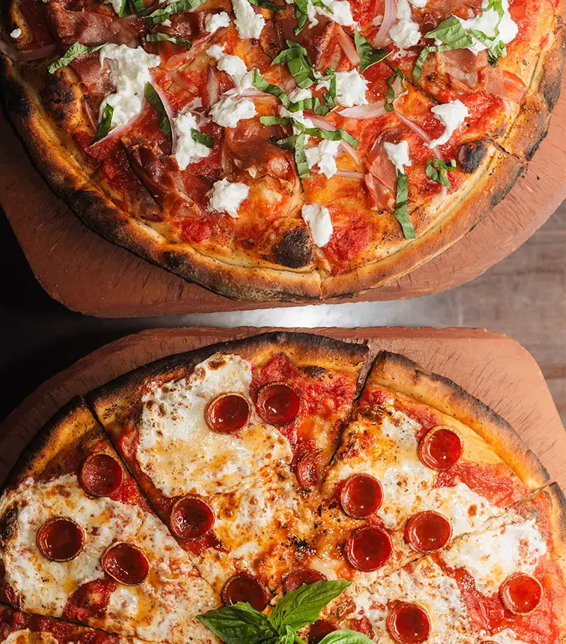 Two freshly baked coal fired pizzas on wooden boards with ricotta, pepperoni, and basil at Anthony’s Italian restaurant kitchen.