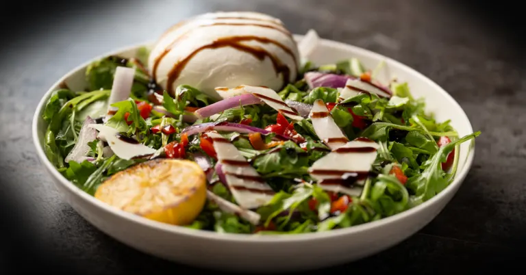 Fresh arugula and creamy burrata topped with tomatoes and onions, served as a vibrant Italian salad at Anthony’s Coal Fired Pizza.