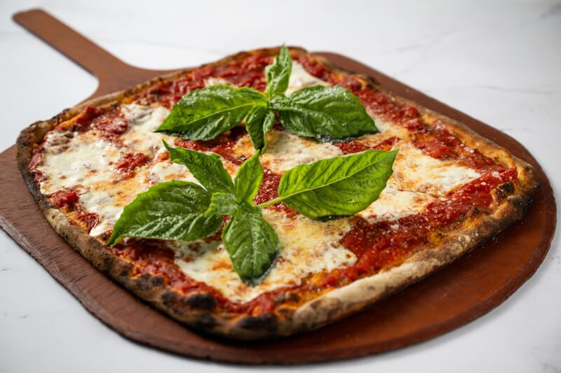 Coal fired square Margherita pizza with fresh mozzarella, rich tomato sauce, and whole basil leaves, served on a crisp, charred crust.