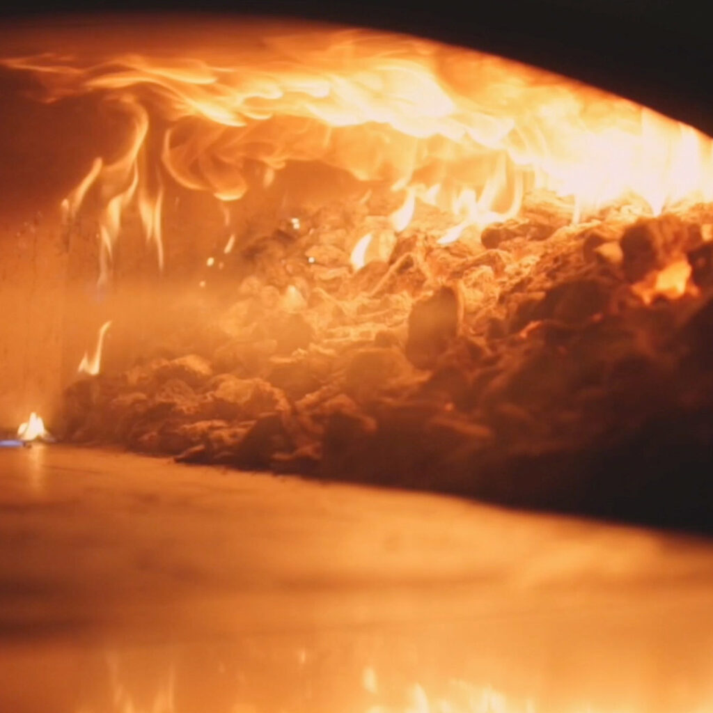Close-up of a wood-fired oven with glowing embers and flames, creating intense heat for baking pizza.