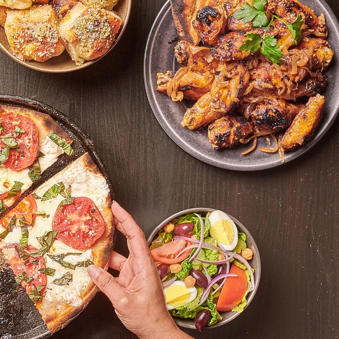 A hand reaching for a pizza slice, surrounded by wings, salad, and bread rolls on a wooden table.